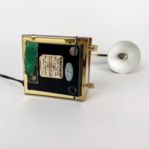 May include: A vintage desk lamp with a rectangular base and a gold-colored frame. The lamp has a small, round, silver-colored shade and a black cord. The base has a label that reads "Model No. 6400".
