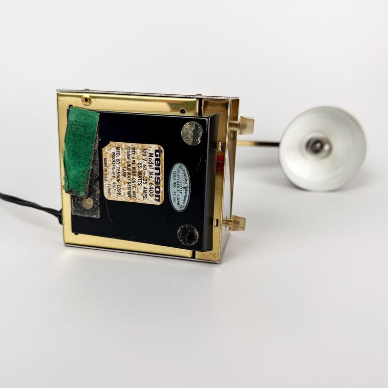 May include: A vintage desk lamp with a rectangular base and a gold-colored frame. The lamp has a small, round, silver-colored shade and a black cord. The base has a label that reads "Model No. 6400".