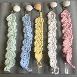 May include: Five pastel-coloured, handmade crochet pacifier clips. The colours are light green, light blue, yellow, white, and pink. Each clip has a metal clasp and a crocheted chain of heart shapes.