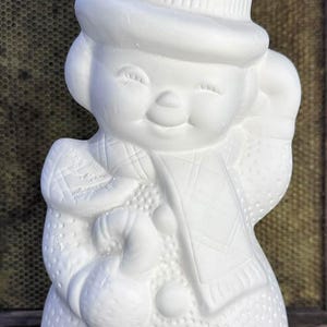 May include: A white ceramic snowman figurine featuring a textured body, a top hat, and a scarf. The snowman is holding a candy cane in one hand and has a smiling expression with closed eyes. The surface of the figurine is matte and unpainted.