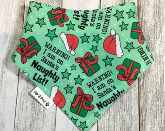May include: A green dog bandana with a festive Christmas design. It features red Santa hats, green gift boxes with red bows, and green stars. The text reads "WARNING! I am on Santa's Naughty List!"