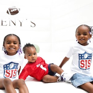 Pode incluir: Tr&ecirc;s crian&ccedil;as vestindo camisas brancas com um logotipo de futebol e o texto "SIS" nelas. A crian&ccedil;a do meio est&aacute; vestindo uma camisa vermelha com o n&uacute;mero "1". As crian&ccedil;as est&atilde;o sentadas em uma superf&iacute;cie branca.