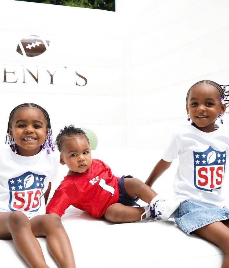 Pode incluir: Tr&ecirc;s crian&ccedil;as vestindo camisas brancas com um logotipo de futebol e o texto "SIS" nelas. A crian&ccedil;a do meio est&aacute; vestindo uma camisa vermelha com o n&uacute;mero "1". As crian&ccedil;as est&atilde;o sentadas em uma superf&iacute;cie branca.