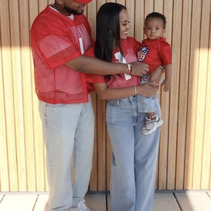 Pode incluir: Uma fam&iacute;lia de tr&ecirc;s pessoas vestindo jerseys de malha vermelha com n&uacute;meros brancos. O pai est&aacute; usando um bon&eacute; de beisebol branco e vermelho com o texto "Daddy's" nele. A m&atilde;e est&aacute; segurando seu beb&ecirc; nos bra&ccedil;os. O beb&ecirc; est&aacute; vestindo um jersey vermelho com o texto "Love" nele. Eles est&atilde;o em p&eacute; na frente de uma parede de madeira.