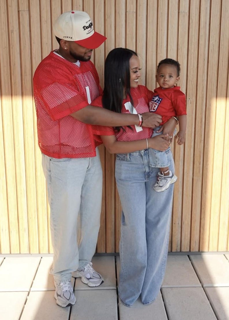 Pode incluir: Uma fam&iacute;lia de tr&ecirc;s pessoas vestindo jerseys de malha vermelha com n&uacute;meros brancos. O pai est&aacute; usando um bon&eacute; de beisebol branco e vermelho com o texto "Daddy's" nele. A m&atilde;e est&aacute; segurando seu beb&ecirc; nos bra&ccedil;os. O beb&ecirc; est&aacute; vestindo um jersey vermelho com o texto "Love" nele. Eles est&atilde;o em p&eacute; na frente de uma parede de madeira.