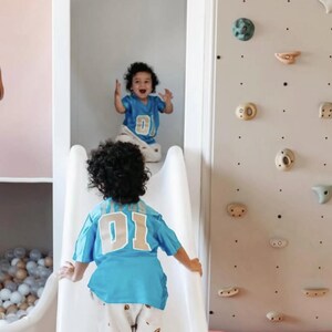 K&ouml;nnte beinhalten: Zwei Kinder mit lockigem Haar tragen passende hellblaue Trikots mit der Nummer 01 in Wei&szlig; und Beige auf dem R&uuml;cken. Ein Kind befindet sich oben auf einer wei&szlig;en Rutsche, das andere unten. Eine Kletterwand mit verschiedenen Griffen ist zu sehen.