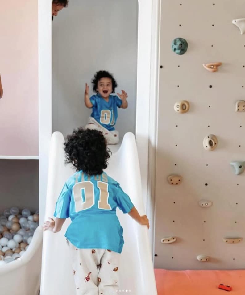 K&ouml;nnte beinhalten: Zwei Kinder mit lockigem Haar tragen passende hellblaue Trikots mit der Nummer 01 in Wei&szlig; und Beige auf dem R&uuml;cken. Ein Kind befindet sich oben auf einer wei&szlig;en Rutsche, das andere unten. Eine Kletterwand mit verschiedenen Griffen ist zu sehen.