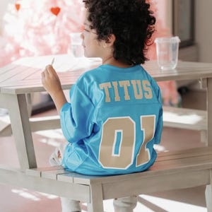 K&ouml;nnte beinhalten: Ein leuchtend blaues Sporttrikot mit dem Namen TITUS in goldenen Blockbuchstaben auf dem oberen R&uuml;cken und der Nummer 01 in gro&szlig;en goldenen Ziffern darunter. Das Trikot besteht aus einem Netzstoff mit kurzen &Auml;rmeln und einem Rundhalsausschnitt.