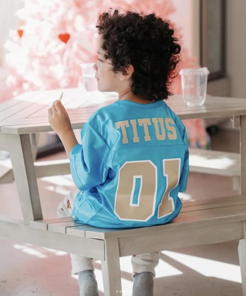 K&ouml;nnte beinhalten: Ein leuchtend blaues Sporttrikot mit dem Namen TITUS in goldenen Blockbuchstaben auf dem oberen R&uuml;cken und der Nummer 01 in gro&szlig;en goldenen Ziffern darunter. Das Trikot besteht aus einem Netzstoff mit kurzen &Auml;rmeln und einem Rundhalsausschnitt.