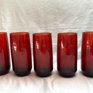 May include: Five tall, red-tinted drinking glasses. Each glass has a dark base and a lighter red upper portion. A subtle etched design is visible on each glass. The glasses are arranged in a row.