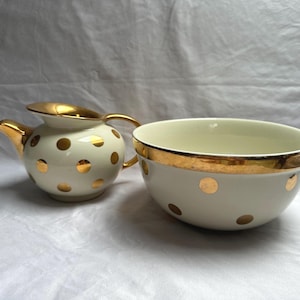 May include: Cream-colored ceramic tea set with gold accents. The set includes a small teapot with a curved spout and handle, and a bowl. Both pieces are decorated with gold polka dots and gold trim.