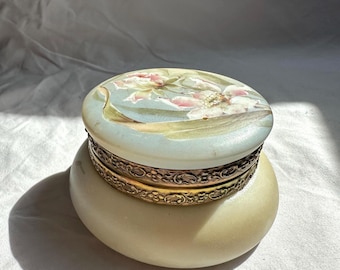 May include: An oval-shaped trinket box with a floral design on the lid. The lid features a light blue background with pink and white flowers. The box has a cream-colored base and a gold-colored band around the middle.