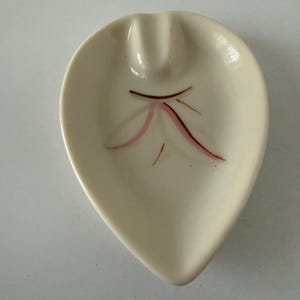 May include: Cream-colored, leaf-shaped ashtray with a decorative design. The ashtray features a central indentation for holding cigarettes and a painted leaf-like design in shades of pink and brown.