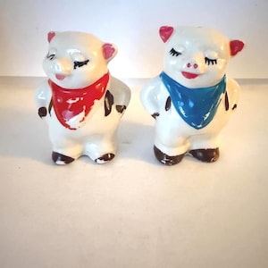 May include: A pair of vintage ceramic pig figurines. The pigs are white with pink ears and red or blue bandanas. They have closed eyes and brown hooves. These decorative figurines are likely salt and pepper shakers.