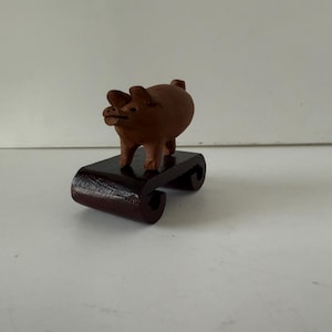 May include: A small, brown wooden pig figurine stands on a dark, curved wooden stand. The pig has a rounded body and small ears. The stand has a glossy finish. The figurine is a decorative item.