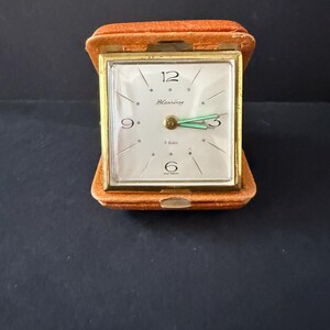 May include: A vintage travel clock in an open, orange case. The clock face is white with gold trim, black numerals, and green hands. The clock face reads "Blessing" and "7 Rubis".
