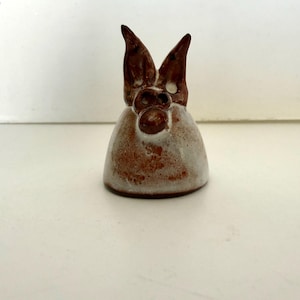 May include: A ceramic figurine of a rabbit with brown ears and a brown nose. The rabbit has a white body with brown speckles. The figurine is sitting on a white surface.