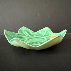 May include: A green ceramic leaf-shaped dish with a textured surface. The dish has a central circular depression and gently curving edges. A small, gold-colored sticker is visible on the rim. The dish is likely for decorative or functional use.