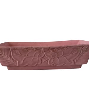 May include: A rectangular, pink ceramic planter with a floral leaf design. The planter has a slightly distressed finish, with a raised base and a wide opening. The overall shape is long and shallow, suitable for various plants.