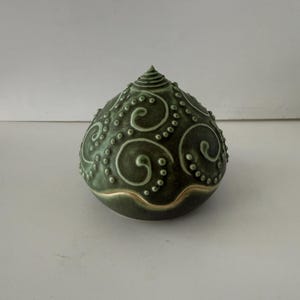 May include: A dark green ceramic decorative object with a rounded shape and a pointed top. The surface features a pattern of swirling designs and small dots, with a gold-colored band at the base. The object is approximately 5 inches tall.