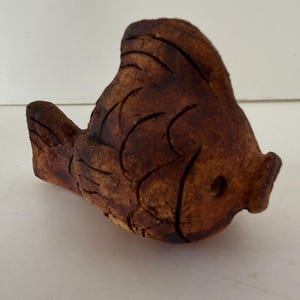 May include: A handcrafted, brown ceramic fish figurine. The fish has carved details to represent scales and fins. The figurine is a warm brown color with darker accents, and a small hole near the mouth.