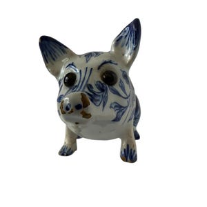 May include: A ceramic pig figurine with a blue and white floral pattern. The pig has large black eyes, a brown snout, and upright ears. The figurine is standing on four small legs.