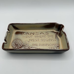 May include: A rectangular ceramic tray with a beige interior and dark brown edges. The tray features an embossed sunflower and the text: KANSAS - OUT WHERE THE WEST BEGINS! THE SUNFLOWER STATE. It has notched corners and a smooth, glazed finish.