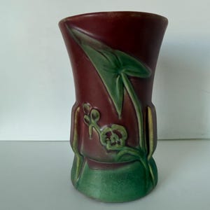 May include: A maroon ceramic vase with a green base and accents. The vase features a raised relief design of a green leaf and flower. The vase is approximately 8 inches tall.
