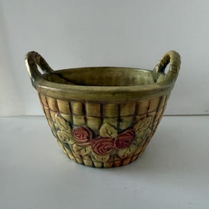 May include: A ceramic basket-shaped planter with two handles. The basket has a woven texture and is painted in shades of yellow, green, and brown. Red roses and green leaves decorate the front of the planter. The handles are a twisted rope design.