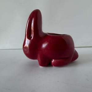May include: A ceramic bunny planter in a deep red color. The bunny has long ears and a small opening on its back for planting small succulents or flowers. The planter is sitting on a white surface.