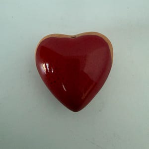 May include: A glossy, heart-shaped decorative object. The heart is a vibrant red color with a light brown border. The object has a smooth, reflective surface, suggesting it is made of ceramic or a similar material. The heart is centered against a plain, light background.