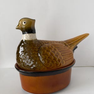 May include: A ceramic pheasant-shaped casserole dish. The dish is primarily brown with a black rim and a detailed pheasant design on the lid. The pheasant has a green head and a white neck ring. A decorative kitchen item.