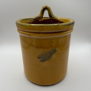May include: A cylindrical mustard-yellow ceramic wall pocket featuring a crackled glaze finish and a single dark cicada illustration on the front. The vessel has a raised rim and a central arched tab at the top for hanging purposes.