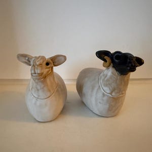 May include: Two ceramic sheep figurines. One is cream-colored with tan ears and face. The other has a black face and horns, with a cream-colored body. Both are in a seated position, with a rounded body and detailed features.