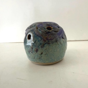May include: A handmade ceramic owl-shaped sculpture with a speckled blue and green glaze. The owl has two eye holes and a beak-like opening. The ceramic piece is a unique decorative item.