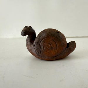 May include: A brown ceramic snail figurine with a textured surface. The snail has a spiral shell design and two small holes on top. The figurine is a decorative item.