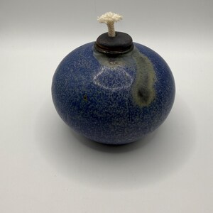 May include: A spherical ceramic oil lamp with a speckled blue glaze and a smooth, dark brown circular cap at the top. A white cotton wick protrudes from the center of the cap. The surface features a subtle gray drip pattern near the opening.