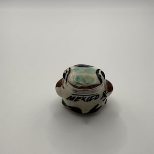 May include: A small, rounded ceramic figurine shaped like a frog with a cream-colored base, brown accents, and black hand-painted markings. The top of the head features a light green patch, and the word MEXICO is written in black script on the front.