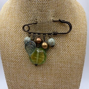 May include: A bronze-toned safety pin brooch featuring a collection of dangling charms. The charms include two round matte bronze beads, two speckled light blue beads, a translucent green leaf-shaped bead, and a round translucent green bead.