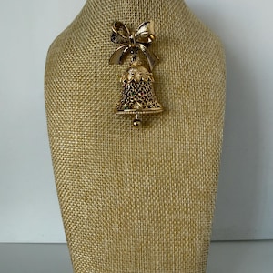 May include: A gold-toned bell-shaped brooch with a bow at the top. The bell has a detailed filigree design with red, green, and clear accents. A small gold ball hangs from the bottom of the bell. The brooch is displayed on a tan mannequin.