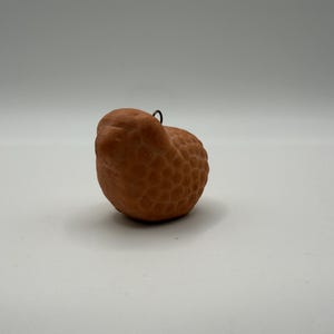 May include: A small, rounded, terracotta-colored ceramic bird figurine with a textured, honeycomb-like surface pattern across its body. A thin, dark metal wire loop is attached to the top of the bird, allowing it to be hung as an ornament.