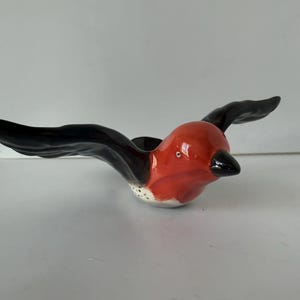 May include: A ceramic bird figurine with outstretched wings. The bird has a red body, black wings, and a black beak. The underside of the bird is white with small black dots. The figurine is designed to appear as if it is in flight.
