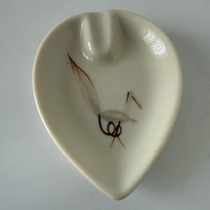 May include: Cream-colored, teardrop-shaped ashtray with a single cigarette rest. The ashtray features a stylized bird design in brown and black. A vintage piece for home decor.