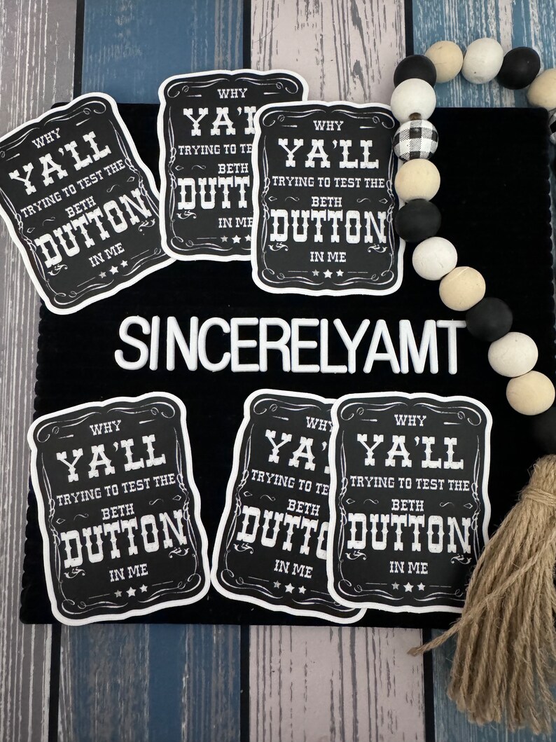 May include: Six black and white stickers with the text "Why Y'all Trying to Test the Beth Dutton in Me" on a black background. The stickers are on a black surface with a white and black beaded garland and a brown tassel.