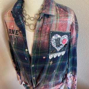 May include: A pink and blue plaid flannel shirt with bleach splatters and a heart-shaped patch sewn on the pocket. The patch has a floral design and a pink button. The shirt has a silver chain necklace on top.