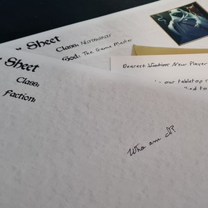 May include: A white sheet of paper with the words "Character Sheet" and "Class: Necromancer" written on it. The sheet is folded in half and there is a gold envelope on top of it. The envelope has the words "Dearest Victim New Player" written on it.