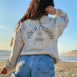 以下が含まれることがあります： 女性は、背中に「YOUR DESIGN here」と書かれた薄いベージュのトレーナーを着ています。彼女は青いデニムのショートパンツを履いており、砂浜に立っています。