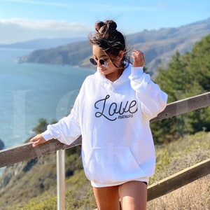 K&ouml;nnte beinhalten: Ein wei&szlig;er, &uuml;bergro&szlig;er Hoodie mit einem schwarzen, kursiv geschriebenen "Love"-Motiv auf der Vorderseite. Der Hoodie wird von einer Person getragen, die auf einem Holzgel&auml;nder steht und auf eine Wasserfl&auml;che blickt.