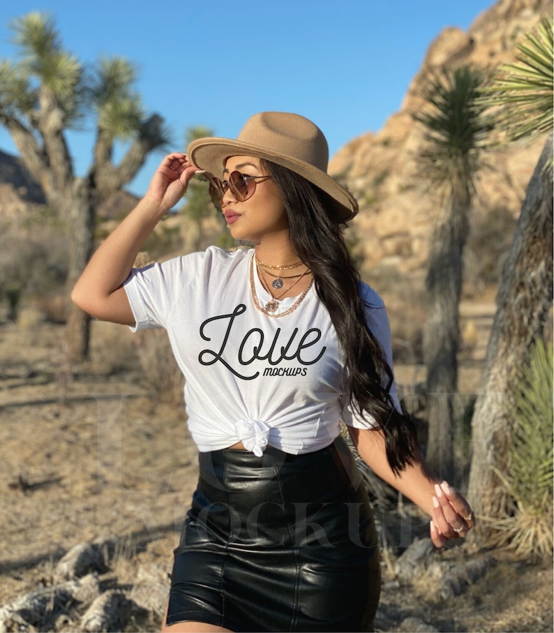 May include: A white t-shirt with the word "Love" in a script font, paired with a black leather skirt. The model is wearing a tan fedora hat and sunglasses. Gold necklaces complete the look. The background features desert vegetation.