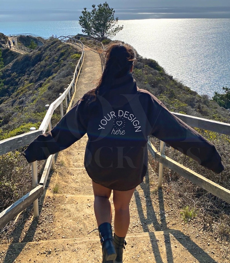 May include: A person wearing a black hoodie with the text "YOUR DESIGN here" on the back, walking down a wooden staircase overlooking a body of water.